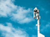 Close-up of a surveillance camera on a pole under a clear blue sky.