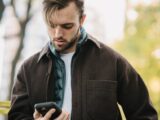 Young bearded man in casual clothes receiving text message on smartphone while standing on street