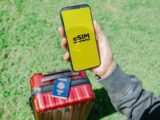 Traveler in Los Angeles holding a smartphone with eSIM near a red suitcase on grass.