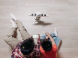 A father and son using a smartphone to control a small humanoid robot on wooden flooring.