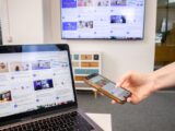 Person using smartphone and laptop in a modern office environment with digital screens.
