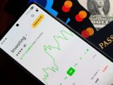 A smartphone showing an investment app with green growth indicators, surrounded by credit cards, US dollars, and a passport.