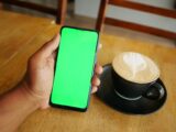 A hand holding a smartphone with green screen beside a latte with art in a cafe.