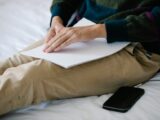 A person using braille on a sheet, seated indoors with a smartphone nearby.