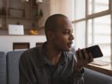 Man holding smartphone and speaking indoors, sitting on sofa by window lighting.