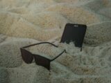 A smartphone and sunglasses partially buried in beach sand, capturing a summer vibe.