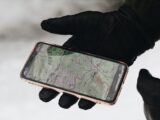 A gloved hand holds a smartphone with a map app displayed, showcasing navigation in cold weather.