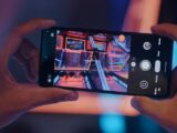 Close-up of hands holding a smartphone, photographing an arcade game screen.