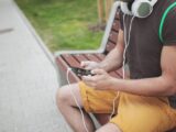 A young adult seated on a park bench, listening to music on headphones connected to a smartphone.
