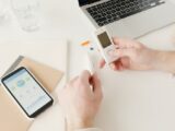 Person checking blood sugar with glucometer on a desk with smartphone and laptop nearby.