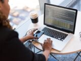 Black woman programming on a laptop with coffee, smartphone, and glasses on a desk in an office.