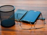 Smartphone, eyeglasses, and wire mesh pen holder on wooden desk.