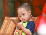 Cute Asian child deeply focused on a smartphone indoors. Captures modern childhood moments.