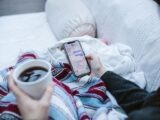 Person relaxing indoors with coffee and smartphone, texting in a cozy setting.