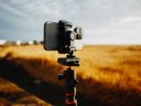 A smartphone is mounted on a tripod capturing the vast golden field under a bright sky.