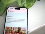 Close-up shot of a smartphone screen showing the OpenAI website with greenery in the background.
