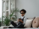 Young adult wearing sunglasses, using a smartphone on a sofa in a modern interior.