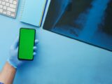 Close-up of a hand in a glove holding a smartphone with a green screen next to x-ray on a blue desk.
