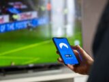 Close-up of a person holding a smartphone with a VPN app, streaming sports on TV.