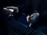 Close-up of hands using smartphone and computer keyboard, holding eyeglasses, focused workspace.