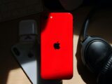 A flat lay of a red iPhone surrounded by AirPods and Bose headphones on a wooden surface.