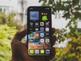 Close-up of a smartphone held outdoors in an autumn setting, showcasing apps and widgets.