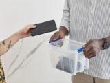 Two adults exchanging a smartphone into a plastic storage box in an indoor setting.