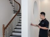 Man adjusting thermostat in stylish home with elegant staircase.