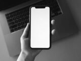 Monochrome image of a hand holding a blank smartphone screen above a laptop, ideal for tech presentations.