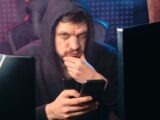 A man in a black hoodie contemplating while using a smartphone, surrounded by digital screens.